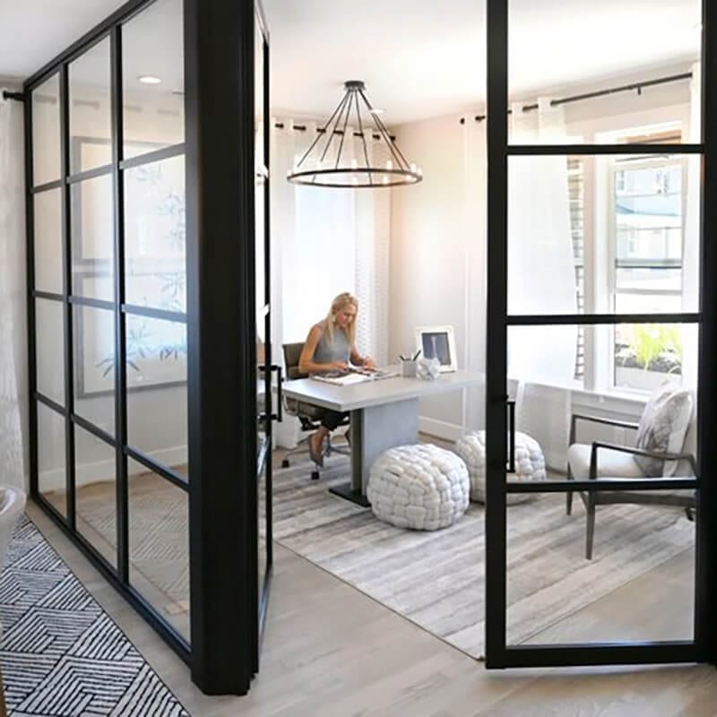 Blonde woman works in home office separated by metal frame clear glass fixed panels and swing doors